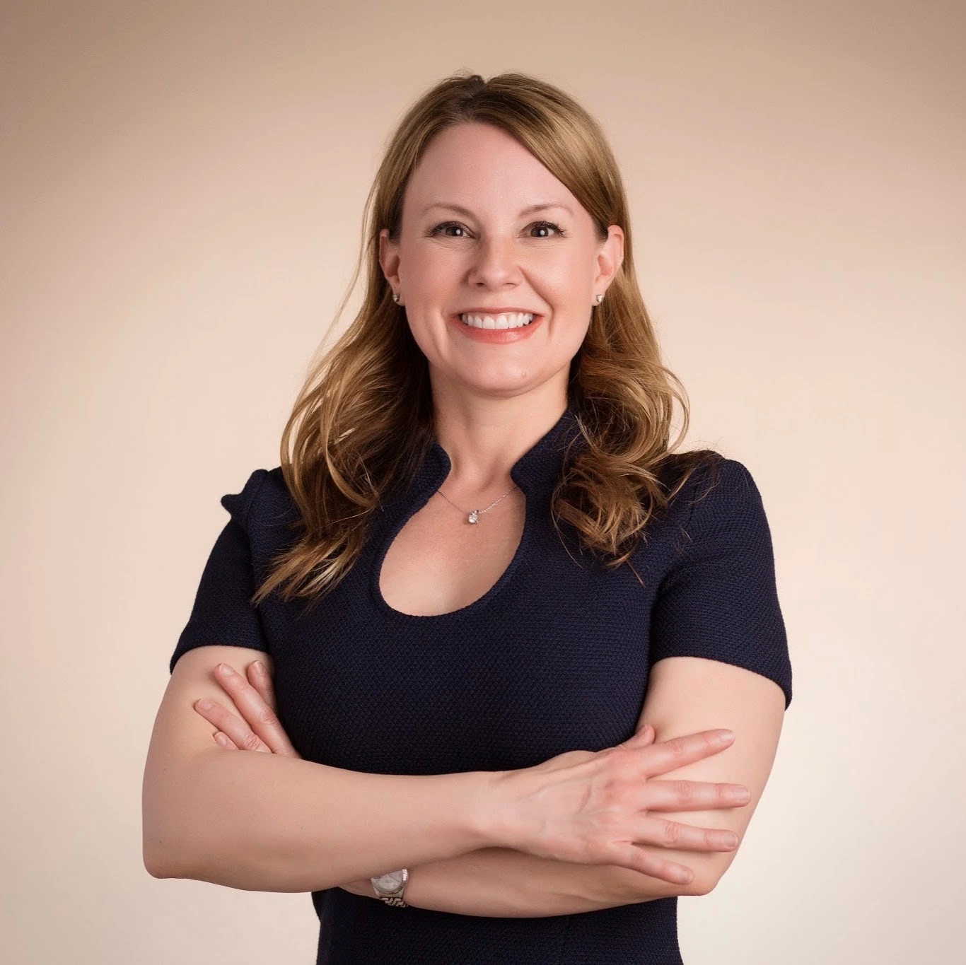 Woman with brown hair smiling at camera, arms crossed, wearing dark blue top against neutral background